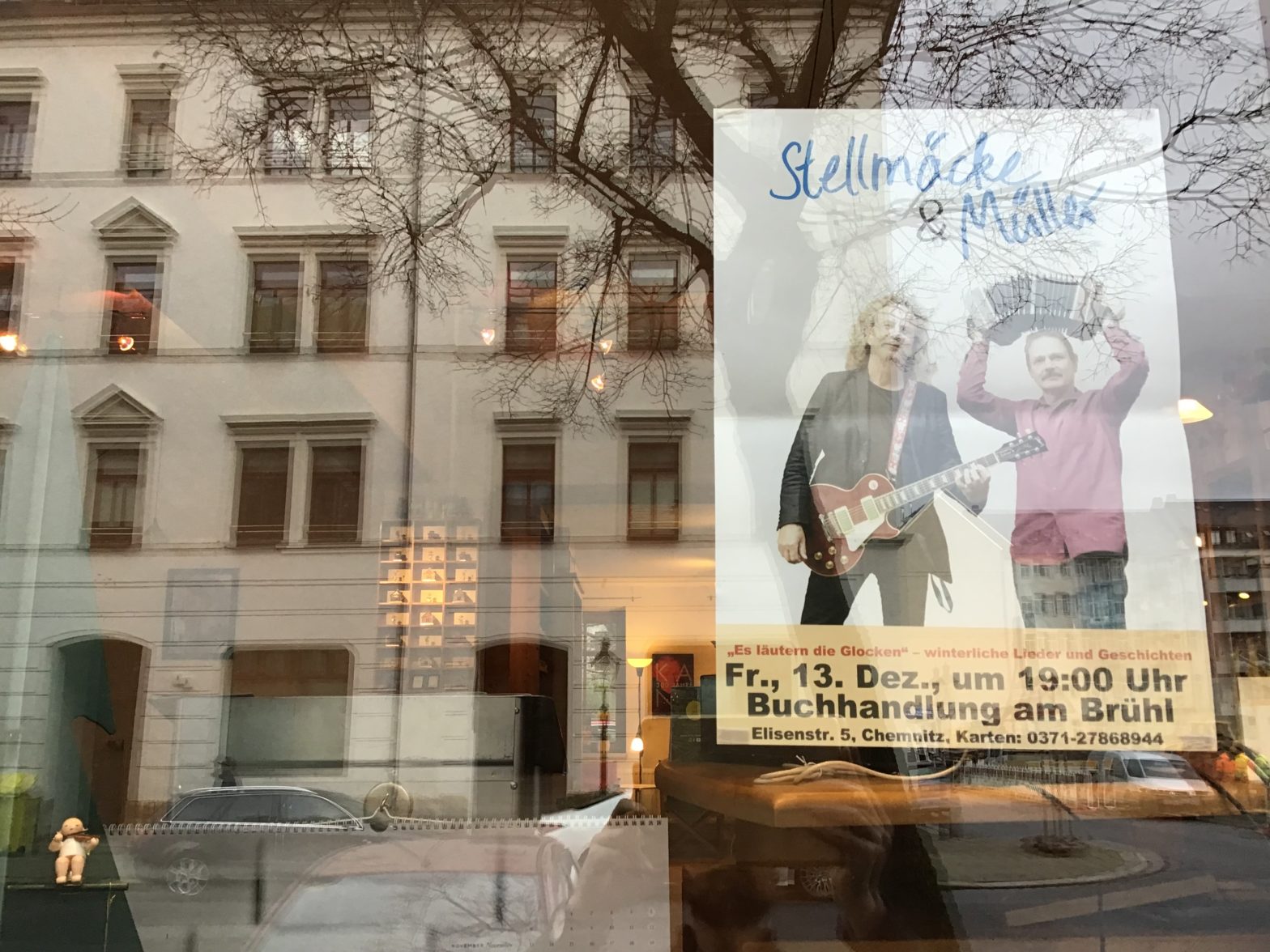 Ein Poster von Stellmäcke & Müller auf dem Schaufenster der Buchhaandlung am Brühl. Im Hintergrund sieht man Teile des Ladeninneren beleuchtet, mit einem Kant-Plakat, Figuren von Wendt & Kühn, Knox Räucherhäsuern und Flade Engeln.