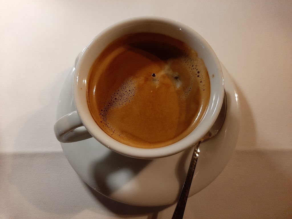 Ein Café Crema in einer kleinen weißen Porzellantasse, die auf einer weißen Untertasse steht. Ein Metalllöffe liegt auf der Untertasse. Der Kaffee ist von oben fotografiert. Im Hintegrund sieht man ein dickes weißes Tischtuch.