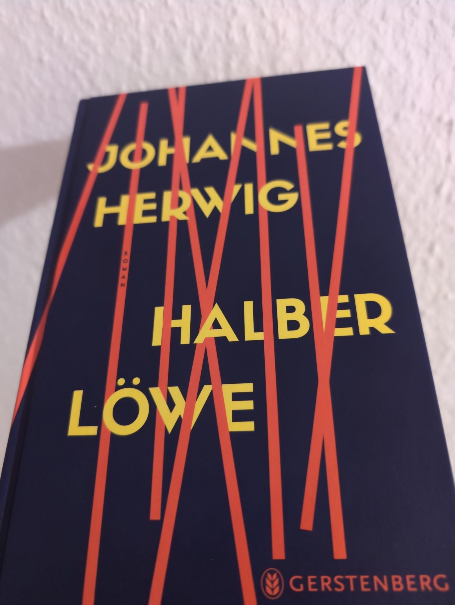 Das Cover von "Halber Löwe" von Johannes Herwig. Orange Streifen verdecken teilweise die gelbe Schrift auf dunkelblauem Grund.