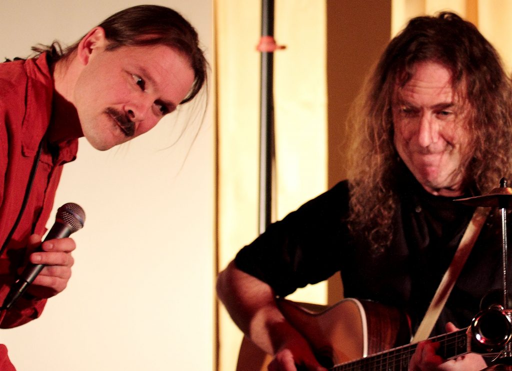 Zu sehen sind Stellmäcke und Müller vor hellem Hintergrund.üller spielt auf der Gitarre und Stellmäcke scheint, mit dem Mikrofon in der Hand, zu lauschen.