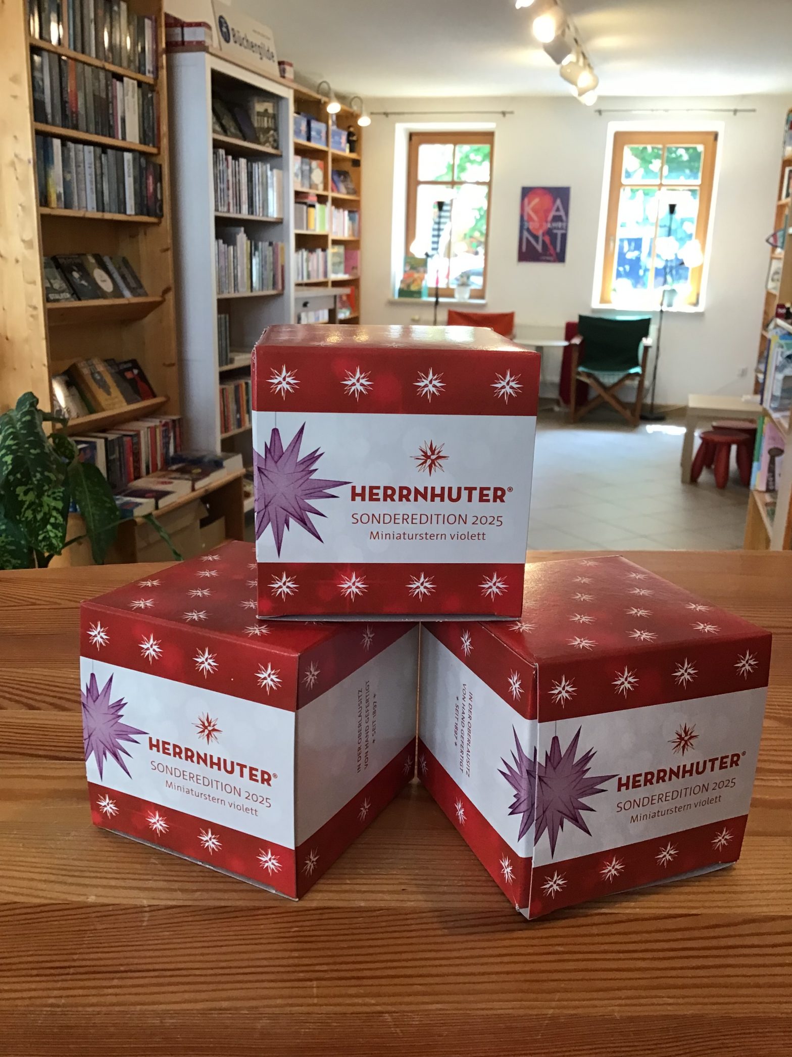 Auf dem Foto sind drei Schachteln der Sonderedition 2025 zu sehen. Im Hintergrund sieht man die Buchhandlung am Brühl.