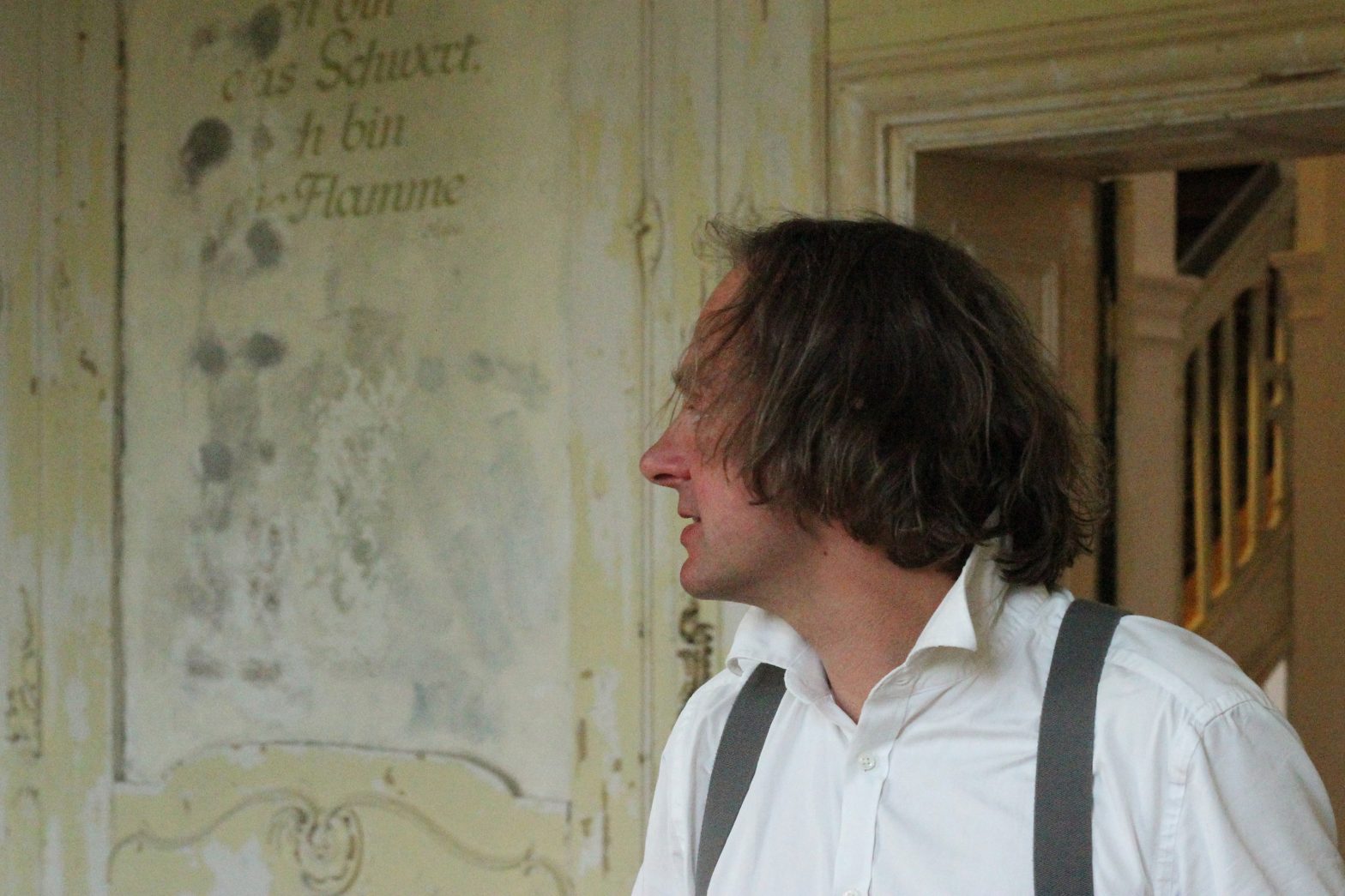 Ein Foto auf dem Tobias Rank zu sehen ist. Man sieht sein Gesicht nur von der Seite, da er in das Linke Off des Fotos blickt. Das Foto scheint in einem alten Haus aufgenommen zu sein Auf der hellen Rückwand stehen verblasste Worte.