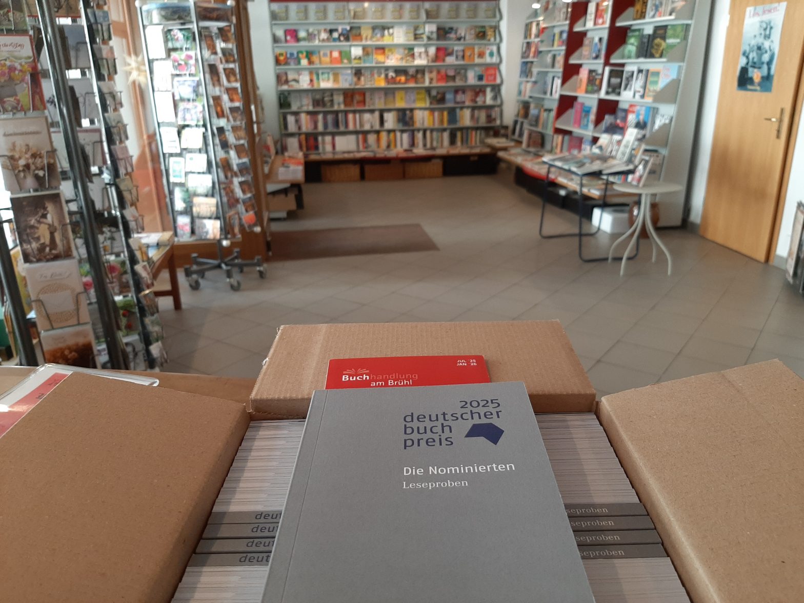 Zu sehen sind die gerade eingetroffenen Leseproben für den Deutschen Buchpreis 2025. Sie befinden sich noch unausgepackt im geöffneten Karton, der auf dem Verkaufstresen der Buchhandlung am Brühl steht. Ein Exemplar liegt auf den anderen, so dass man es frontal sehen kann. Es ist ein graues Taschenbuch, auf dem in dunkelblauer Schrift "2025 Deutscher Buchpreis" steht und darunter, etwas kleiner, in weiß: "Die Nominierten". Und wieder darunter, wieder etwas kleiner und ebenfalls in weißer Schrift: "Leseproben". Im Hintergrund ist die Romanwand der Buchhandlung am Brühl zu sehen.