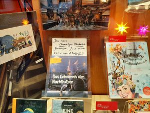 Zu sehen ist eines der Schaufenster der Buchhandlung am Brühl, im Zentrum das Chemnitzer Märchenbuch "Das Geheimnis der Nachtkatzen" der Edition Claus, das bei uns in der Buchhandlung am Brühl vorrätig und noch bis zum 22.12.25 3x zu verlosen ist.